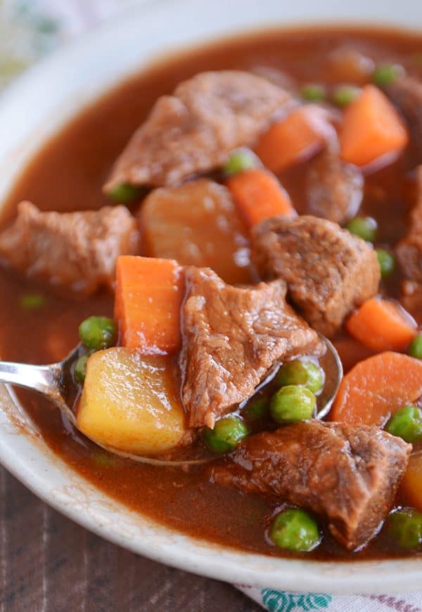 A bowl full of stew filled with cubes of beef, carrots, peas, and potatoes with a spoon taking a bite out.