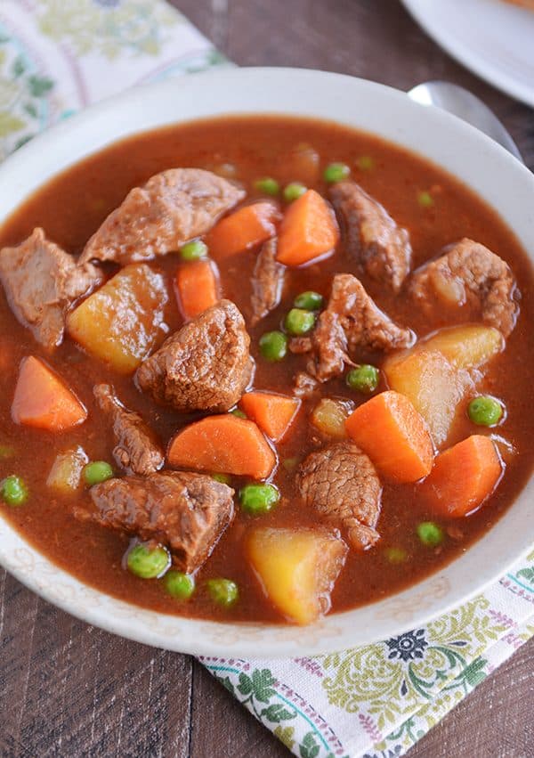 A bowl full of stew filled with cubes of beef, carrots, peas, and potatoes. 