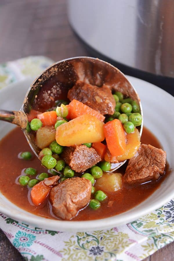 A ladle full of beef, potatoes, carrots, peas, and stew broth being poured into a bowl.