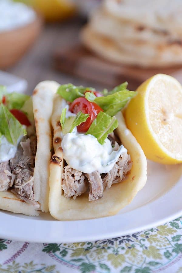 A white plate of two Greek pork tacos topped with lettuce and tomatoes with a half lemon on the side. 