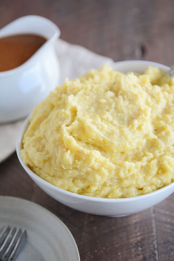 Large bowl full of mashed potatoes, with a gravy boat of brown gravy next to it. 