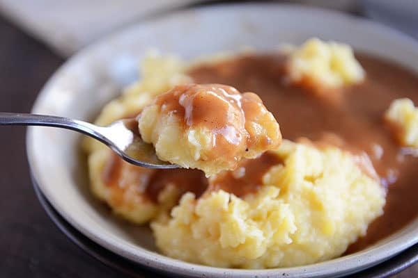 A spoonful of mashed potatoes and brown gravy, with a bowl full of mashed potatoes and brown gravy in the background. 