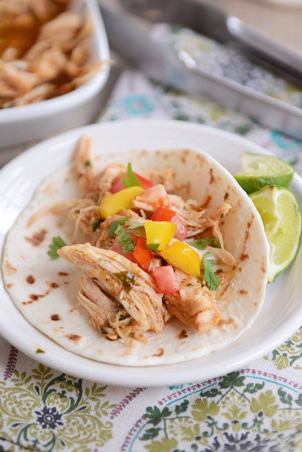 A tortilla filled with cooked, shredded chicken, chunks of pepper and tomato, and 2 lime slices on the side of the tortilla. 
