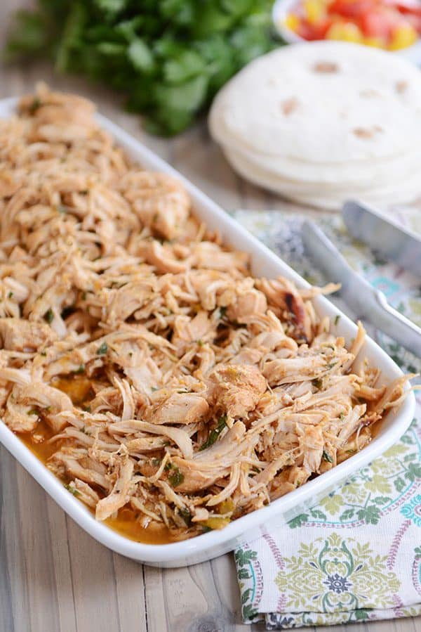 A white dish full of cooked, shredded chicken with bits of cilantro in it and a stack of tortillas and a bunch of cilantro in the background.