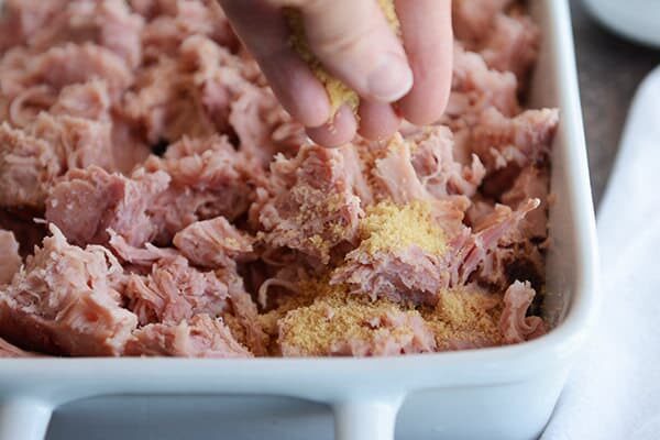 A white dish full of shredded, baked ham with seasoning being sprinkled on the top.