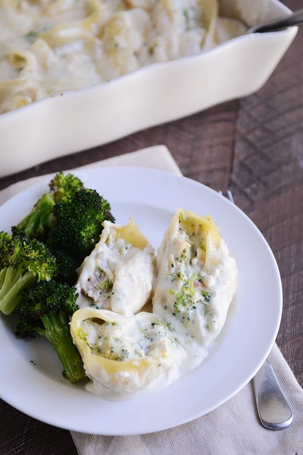 A white plate with three jumbo stuffed alfredo pasta shells next to a helping of cooked broccoli.