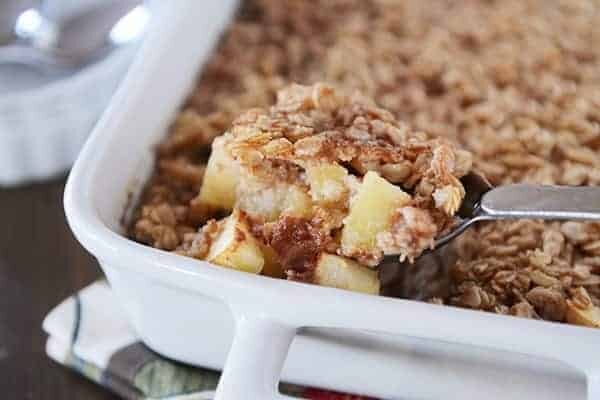 A spoon taking a bite full of oatmeal with chunks of apple, out of a white dish. 