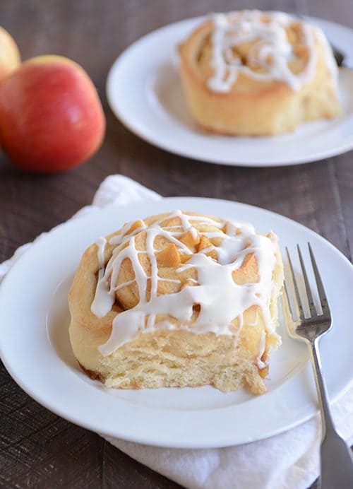 Two icing drizzled apple pie cinnamon rolls on white plates.