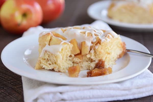 A cinnamon roll with chunks of apple and frosting and a bite taken out on a white plate.