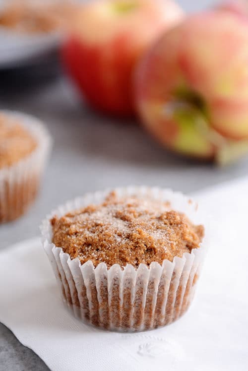 A sugar-dusted muffin in a white liner with apples in the background.