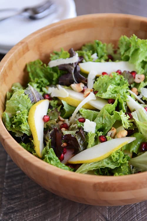 A wooden salad bowl full of a green salad topped with sliced pears, nuts, and shredded parmesan.