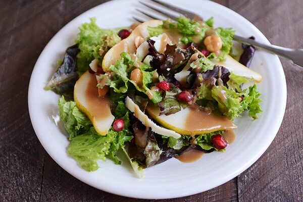A white plate with green salad topped with sliced pears, pomegranates, and dressing.
