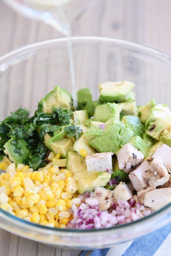 Pouring lemony dressing over avocado chicken salad in glass bowl.