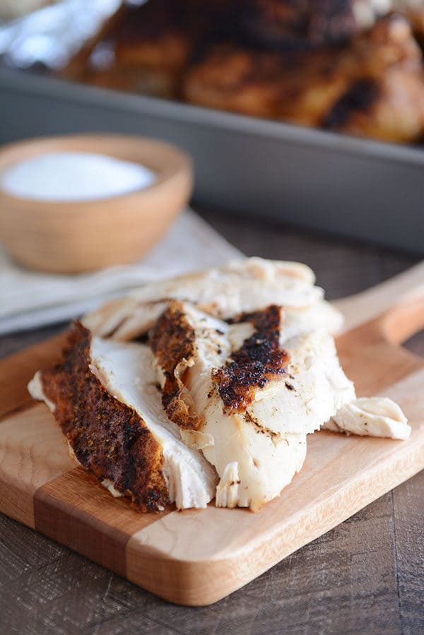 Big pieces of honey roast chicken on a wooden cutting board with a small bowl of salt beside it.