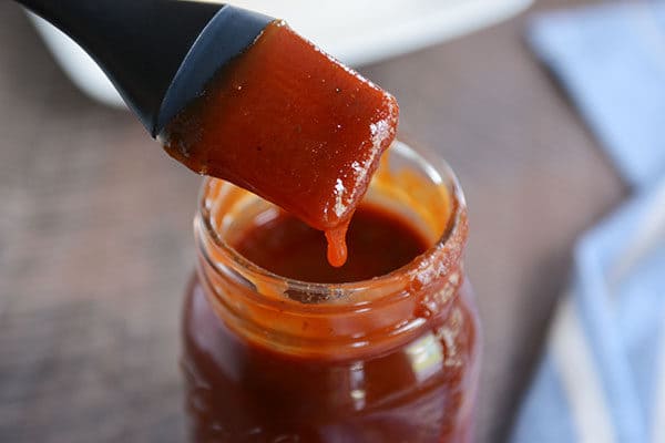 A mason jar full of BBQ sauce, with a drip coming down the side and a pastry brush with sauce over the top. 