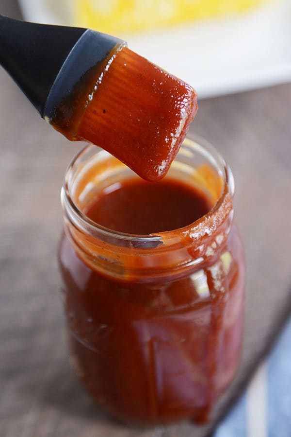 A mason jar full of BBQ sauce, with a drip coming down the side and a pastry brush with sauce over the top. 