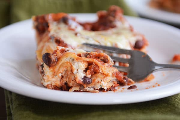 A square of cheesy black bean lasagna on a white plate with a fork taking a bite out. 