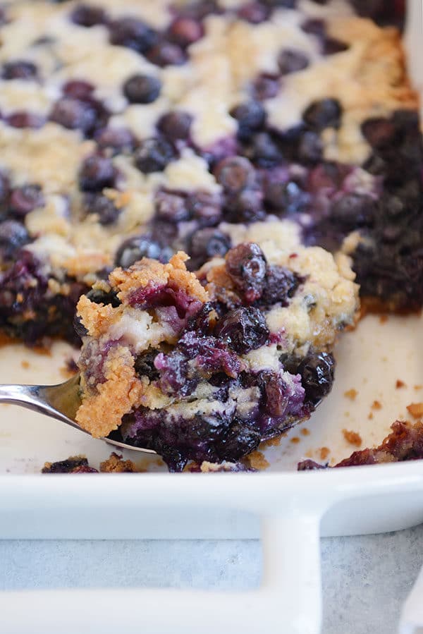 A large metal spoon taking a big scoop of blueberry cake out of a white cake dish.