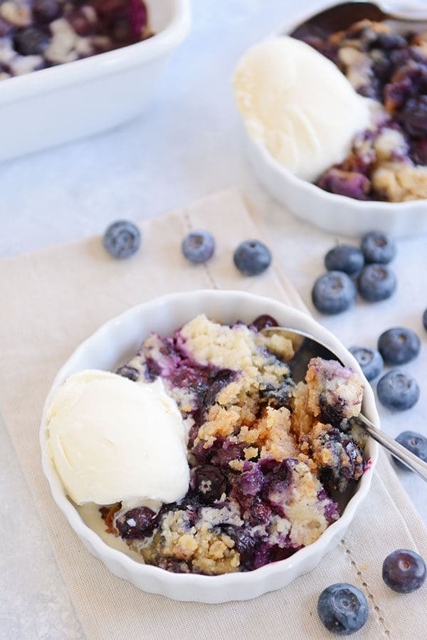 White ramekins of blueberry cake with vanilla ice cream on the side and some sprinkled fresh blueberries on the side.