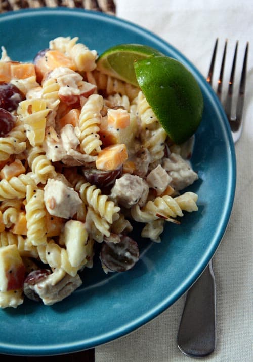 Blue bowl full of creamy chicken salad pasta with cubes of cheese and sliced grapes and two lime wedges on the side of the bowl.