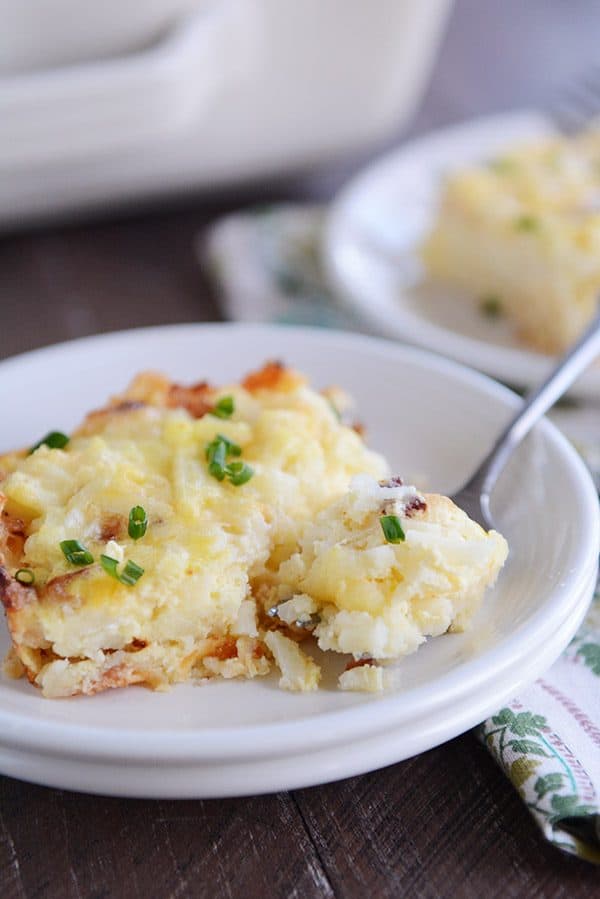 A piece of cheesy egg and hash brown casserole on a white plate, with a fork taking a bite out.