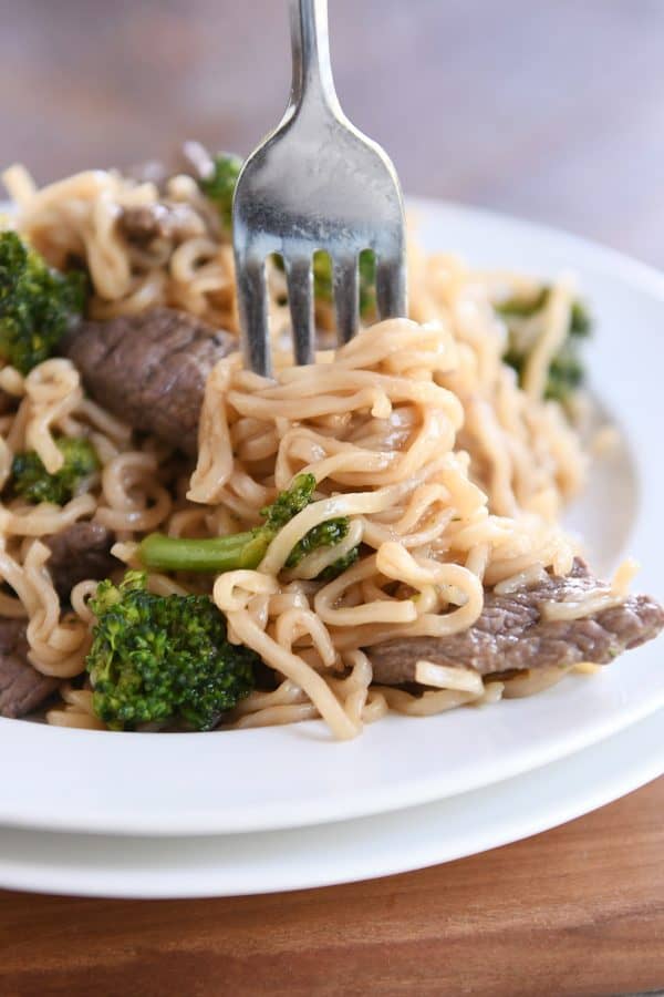 Twisting ramen noodles on fork with beef and broccoli on white plate.