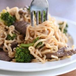 Twisting ramen noodles on fork with beef and broccoli on white plate.