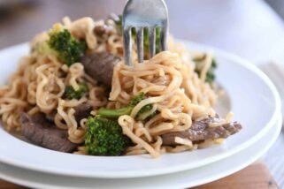 Twisting ramen noodles on fork with beef and broccoli on white plate.