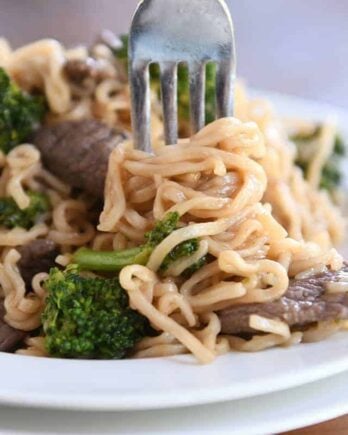 Twisting ramen noodles on fork with beef and broccoli on white plate.
