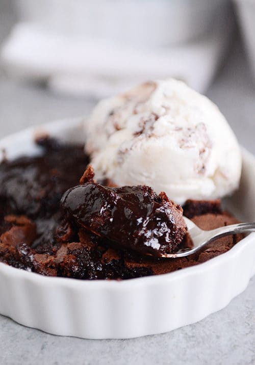 A white ramekin full of brownie pudding, with a scoop of ice cream on the side, and a spoon taking a bite out.