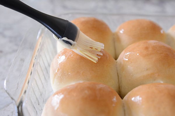 Baked French bread rolls in a glass pan getting brushed with butter. 