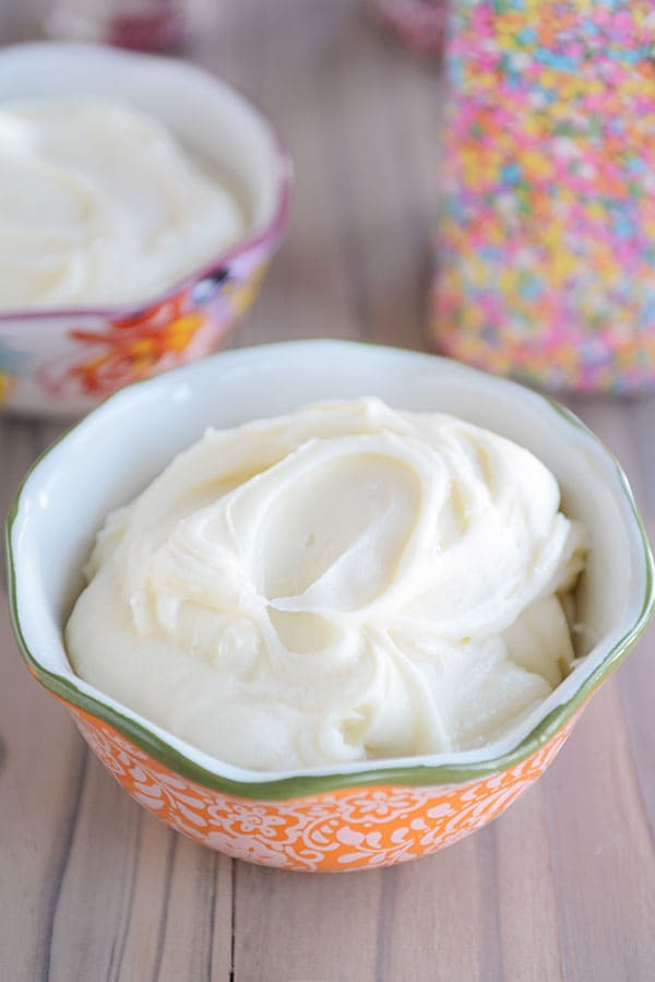 A bowl full of vanilla buttercream frosting with a jar of sprinkles and another bowl of frosting in the background.