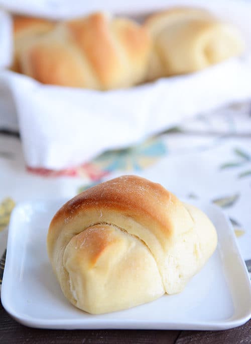 A golden brown roll on a white plate with a basket of rolls behind it.