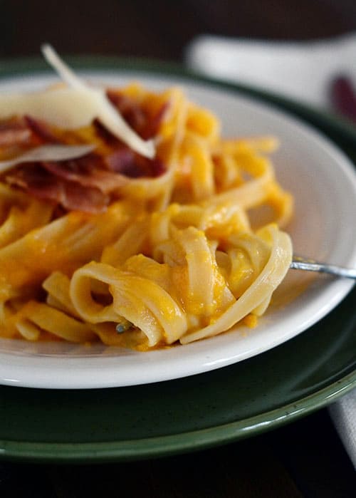 A white plate with cooked linguine pasta topped with bacon and parmesan cheese.