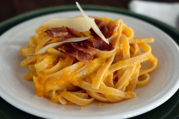 A white plate of butternut squash pasta with chopped bacon and parmesan on top.