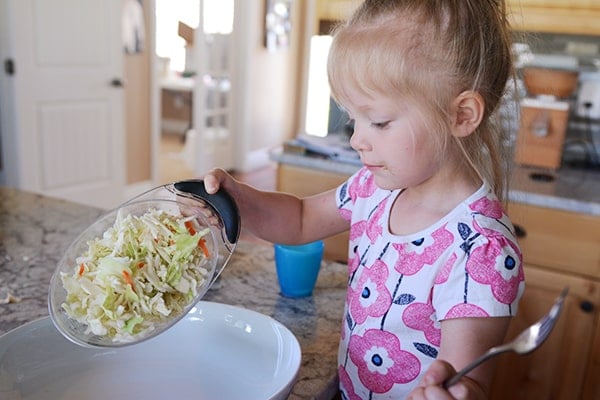 A little girl pouring shredded cabbage into a large white platter. 