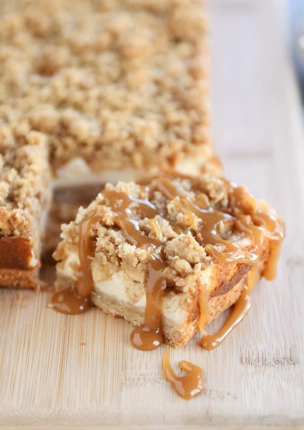 Slice of caramel apple cheesecake bars drizzled with caramel on wood cutting board.