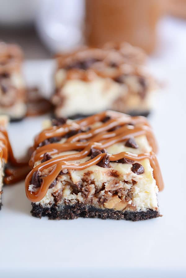Squares of caramel drizzled chocolate chip cheesecake bars with an Oreo crust on a white countertop.