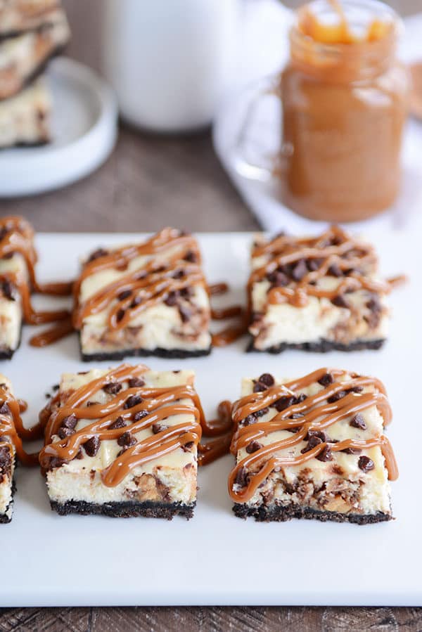 Snickers Oreo cheesecake bars topped with chocolate chips and caramel drizzle on a white platter.