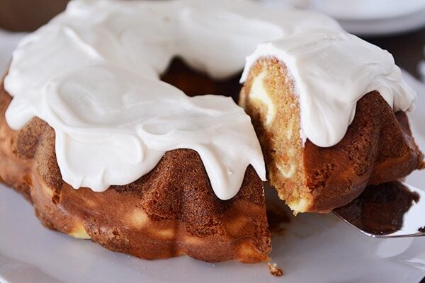 A frosted carrot cake and cheesecake swirled bundt cake getting a piece lifted out of it.