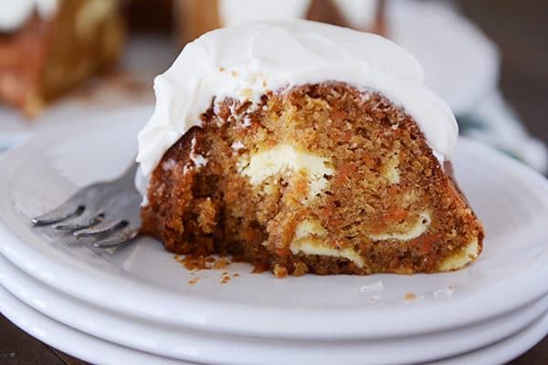 A piece of swirled carrot cheesecake bundt cake with frosting on a stack of white plates.