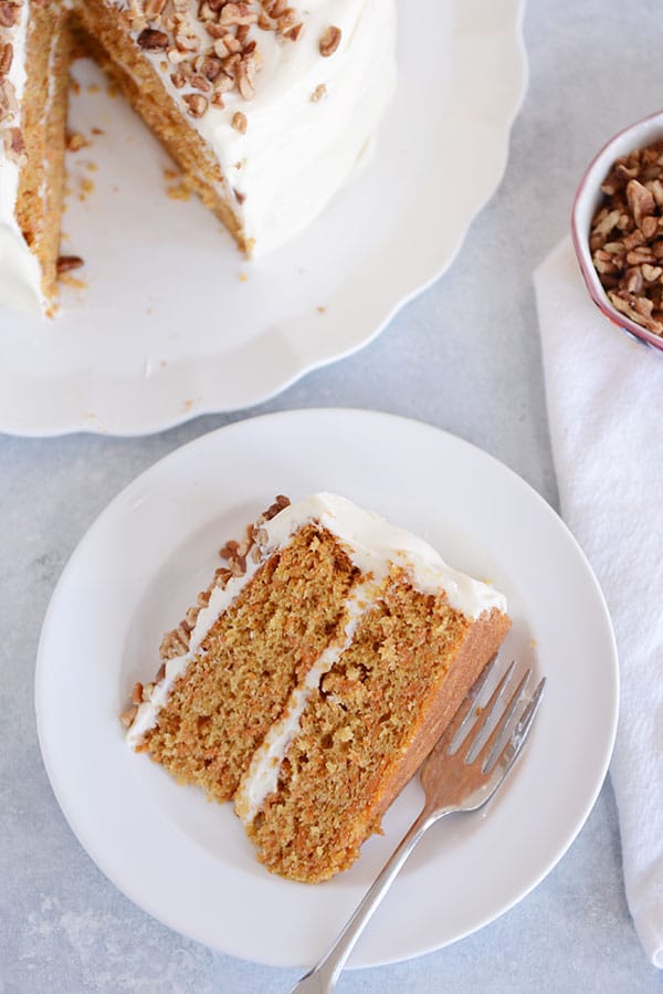 A top view of a slice of carrot cake with two layers, white frosting in the middle, and chopped nuts on top on a plate, and the rest of the cake in the background.