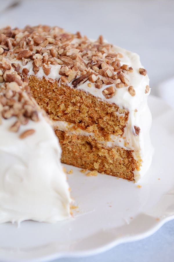 A white-frosted, nut-topped carrot cake with a slice taken out on a white cake plate.