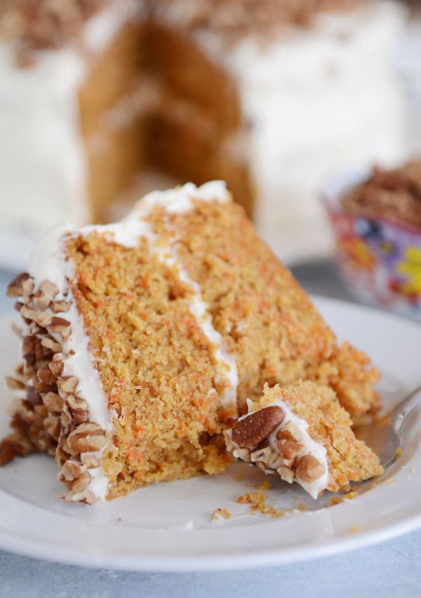 A slice of carrot cake with two layers, white frosting in the middle, and chopped nuts on top on a plate, and the rest of the cake in the background.