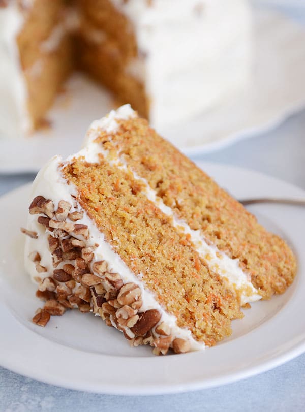 A plate of carrot cake with two layers, white frosting in the middle, and chopped nuts on top, and the rest of the cake in the background. 