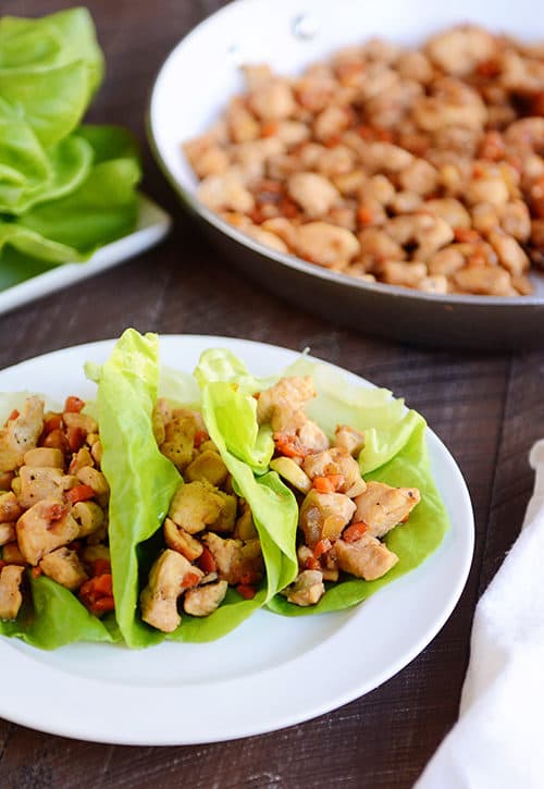 Three cashew chicken lettuce wraps on a white plate with a skillet of chicken mixture behind it.