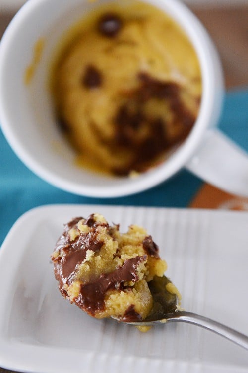 A mug full of a baked chocolate chip mug cookie, with one bite taken out on a spoon in the front of the mug. 