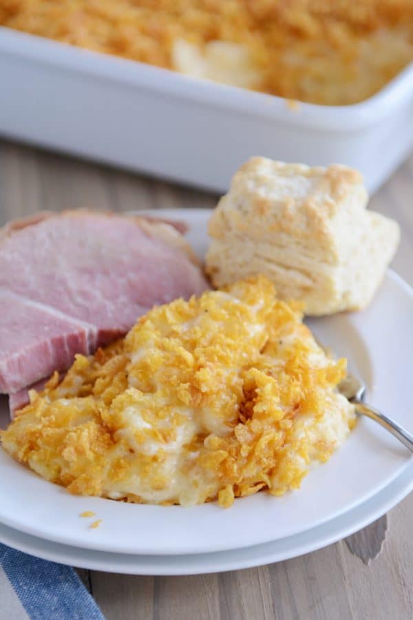 Scoop of cheesy funeral potatoes on plate with ham and biscuit.