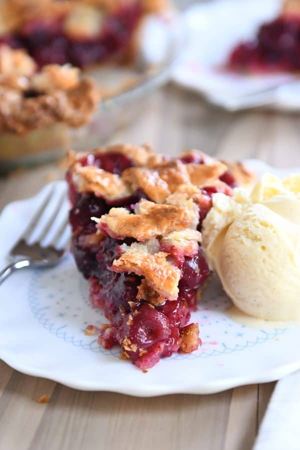 Piece of cherry pie on white plate with scoop of vanilla ice cream.