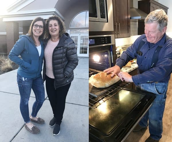 Two woman standing side by side next to a picture of a man pulling bread out of the oven.